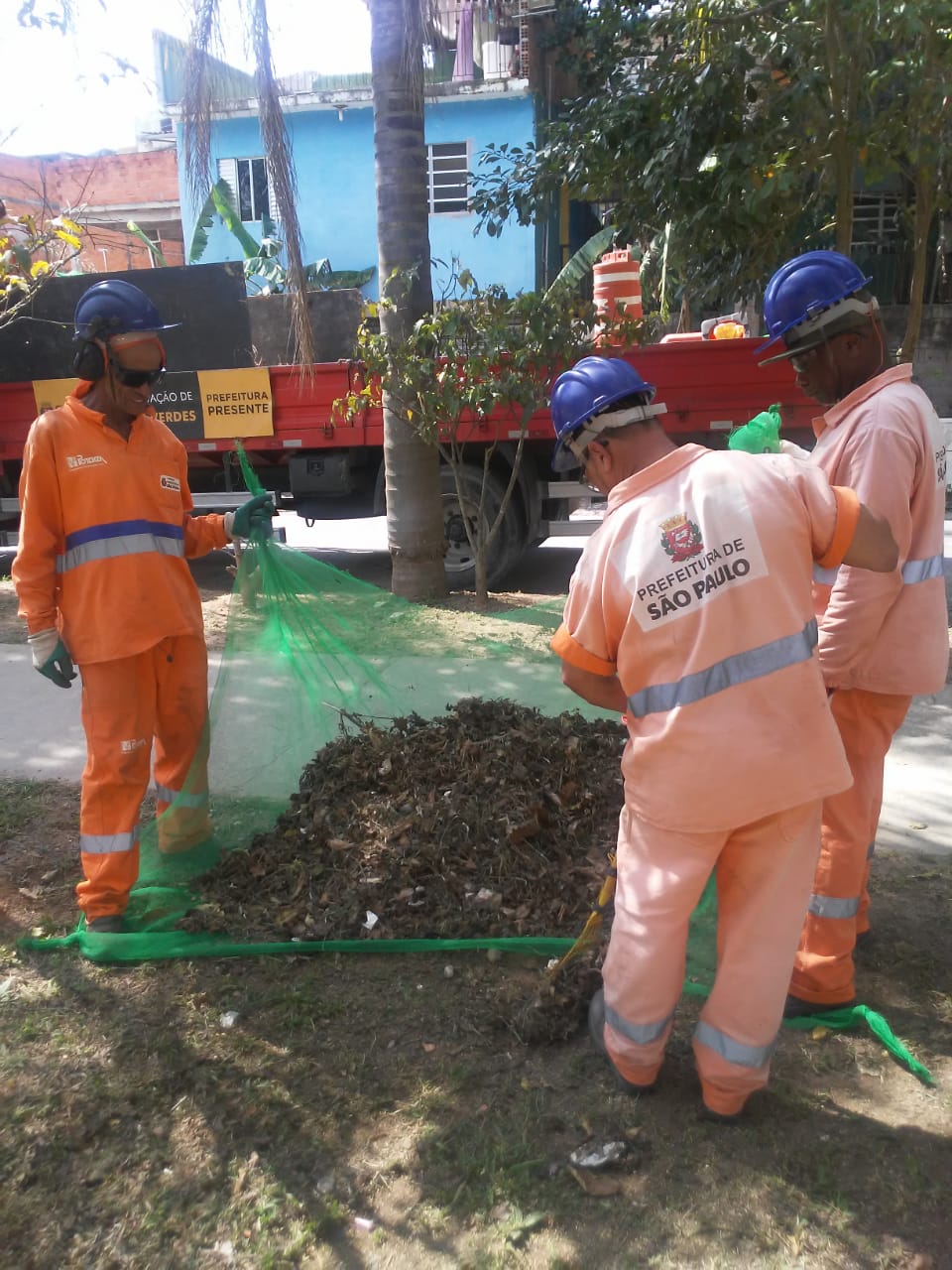 Três funcionários de uniformes uniformes laranjas e capacetes azuis seguram uma rede com resíduos de grama.